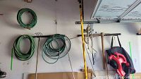 Wall-mounted coiled green garden hoses, some garden tools including rakes, and a black and red leaf blower hanging with its hose attachment.
