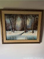 Framed winter landscape painting signed by J. McCurdy showing trees, snow, and reflecting water in neutral colored frame.