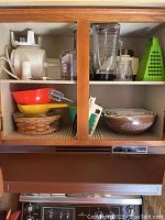 Wide view of upper kitchen cabinet showing the Sunbeam mixer, clear plastic food processor, Pyrex bowls in multiple colors, and a green plastic grater.