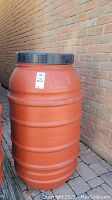 Full view of orange cylindrical rain barrel with black lid, outdoor setting, showing size and shape.