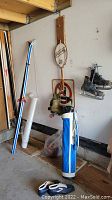 Photo showing blue and white vintage golf bag with golf clubs, black CCM hockey skates hanging on wall, a vintage roller skate and white blue cross country ski shoes on floor.