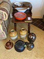Photo showing wooden side table with two shelves and multiple ceramic and stoneware items arranged on and around it including platters, bowls, and jugs.
