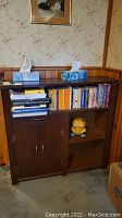 Front view of vintage wooden cabinet with shelves containing books and a small toy, placed against a wood-paneled wall.