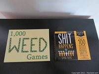 Two game boxes side by side: 1,000 Weed Games (green and beige box) and Shit Happens Card Game (black and yellow box).