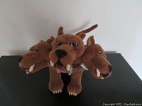 Front and side view of brown three-headed dog plush toy with silver collar spikes, showing all three heads and body.