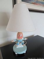 Full view of the Humpty Dumpty wooden lamp with white conical lampshade and light bulb installed, showing the figure's face and blue painted base with four white legs.