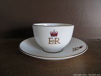 White Minton fine bone china teacup and saucer featuring Queen Elizabeth II insignia with gold and red crown detail, photographed on dark surface and neutral wall.