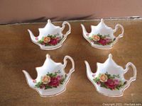 Four white ceramic tea bag trays shaped like teapots with rose floral decoration and gold edge trim set on a flat surface.