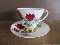 Bone china teacup and saucer with red rose design, showing handle and gold trim on base and rim with some wear.