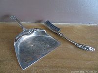 Top view showing silver plated crumb dustpan and matching silver plated crumb brush with floral decorations and black bristles, both items placed on a table.