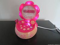 Front view of the 2007 talking Barbie alarm clock showing the digital red LED display with '12:00', pink flower-shaped bezel and decorative butterfly on pink and cream base.