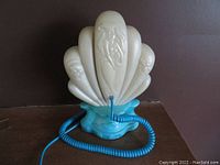 Front view showing off-white seashell handset with character reliefs and turquoise decorative base with coiled phone cord.