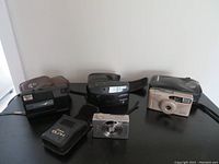 Four vintage cameras shown together: Minolta Freedom Memory Maker II with case (front, brown), Canon Elph 2 with black case, Vivitar 830PZ beige camera, Kodak Tele Disc camera all displayed on black surface.