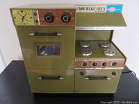 Front view of olive green Easy-Bake Oven showing clock, knobs, oven window, lower compartments and cooktop