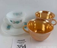 Three cups and one saucer: one white opaque glass cup and saucer set with blue floral pattern (Blue Carnation), two peach luster teacups with shiny metallic finish.