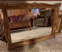 Front view showing the large rectangular mirror with gold ornate frame and detailed scrollwork, reflecting furniture and carpet.