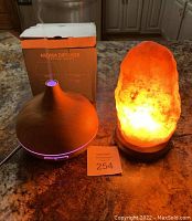 Photo shows a lit Himalayan salt lamp with bright orange glow and an aroma diffuser with purple LED light on a granite countertop, with diffuser box in background.