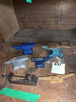 Four anvils in different colors and sizes with a metal clamp and wooden plane handle on a wooden surface.