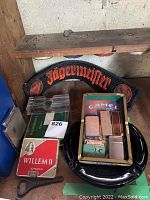 Photo showing Jagermeister rubber bar runner, ashtrays, cigarette packs, and assorted lighters in an box with a bottle opener.