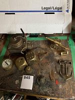 Overview of multiple brass items including large dice, brass anchor, cannon model, dial gauge, and Marsh instrument on a workbench.