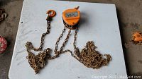Orange ROK chain hoist with attached metal chains and hooks laid out on white surface showing overall condition.