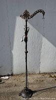 Full view of tall metal floor lamp with black base standing outside against white wall. Cord and plug visible wrapped around the pole.