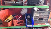 Shelf with orange metal organizer box, blue plastic bin, small plastic drawer unit and gauges including one cracked dial gauge