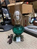 Full view of the vintage turquoise glass lamp with brass base and glass chimney, sitting unplugged on carpeted floor with boxes and clutter in background.