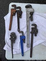 Seven metal pipe wrenches of different sizes and brands laid out on a white towel outdoors, showing various degrees of rust and wear.