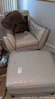 Beige leather armchair and matching ottoman wrapped in clear protective plastic with dark brown toss pillow and blanket on the armchair's left arm.