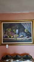 Framed painting on canvas showing various grapes, a basket, and an urn on a table against a muted background.