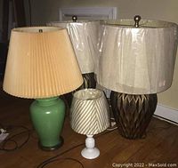 Four lamps of varying sizes with lampshades, two large dark brown lamps with beige shades wrapped in plastic, one medium green lamp with pleated beige shade, and one small white lamp with striped shade wrapped in plastic.