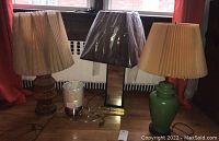 Four assorted table lamps with shades grouped on floor by window featuring wooden carved base lamp, rectangular brown base lamp, green ceramic base lamp, and a small modern lamp with metal base and white shade.