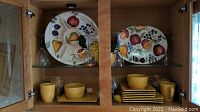 Two large ceramic serving platters with fruit designs on upper wooden cabinet shelves, yellow bowls, and square plates below, clear stemmed glasses around.