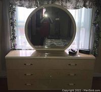Front view of the dresser showing six drawers and the large round mirror attached on top. Light cream color with gold-tone drawer handles. Some wear and scuff marks present.