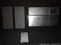 Two silver Panasonic speakers with visible stains alongside a Panasonic silver CD stereo system with buttons and display panel under low light.