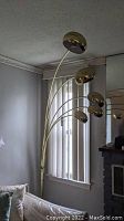 Full view of the five-arm arch floor lamp standing next to a window and sofa, showing the lamp's height and gold finish overall.