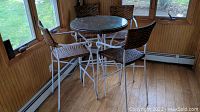 Bar-height patio set with four wicker wrapped chairs surrounding a square wicker topped table with glass top, white painted metal frame, placed indoors near windows.