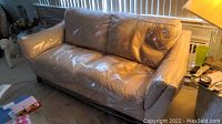 Beige leather sofa covered by clear plastic, showing overall condition and shape of sofa in typical living room environment.