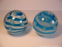 Two Murano style glass paperweights with blue twist canes over white lace ground and bubbles, shown side by side on white background.