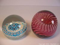Two Murano glass paperweights shown side by side on a flat surface. One has a blue and white concentric Millefiori pattern and the other a pink swirl design.