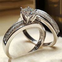 Close up photo of two rings showing the engagement ring with a large simulated white sapphire center and smaller stones on band; wedding band similarly adorned with smaller simulated stones.