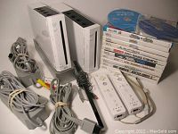 Two white Wii consoles with stands alongside cables, remotes, sensor bar and stacked game cases with discs on top