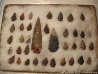 Full view of 35 two-notched arrowheads displayed on white padding with two large spearheads in the center.