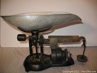 Front view of black cast iron balance scale featuring large aluminum bowl pan, weighted balance arm with sliding weight, and hanging weight holder with attached weight.