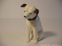 Front left profile of the porcelain Nipper dog figurine showing the dog seated, painted brown ears, and studded collar.