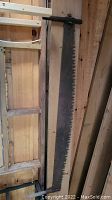 Full length photo of the large vintage two man crosscut saw hanging vertically on a wooden wall, showing metal blade with large teeth and aged wooden handles.