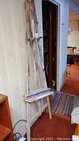 Full view of wooden artist easel leaning against wall with paint splatters on wood and adjustable tray.