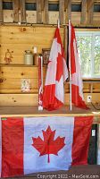 Five Canadian flags, three attached to white poles, displayed against a wooden wall, with two additional flags with symbols partially visible wrapped in sleeves.