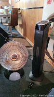 Photo of Presto Heat Dish parabolic heater and Noma tower heater placed side by side on the floor with a brown chair and wooden panel wall in the background.